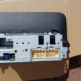 Rear view of a car stereo head unit on a cardboard surface, showing its metal backplate with multiple connectors (gold multi-pin harness, small blue connector, and other ports) and mounting screws.